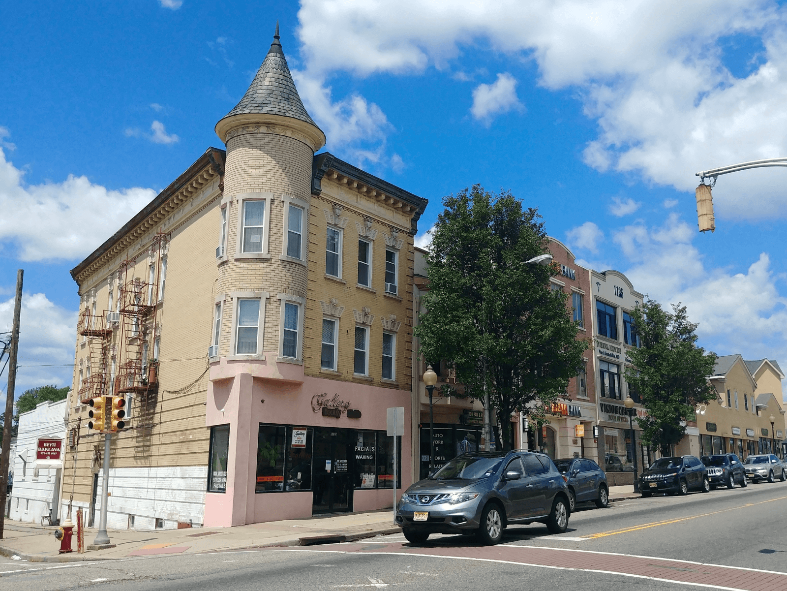 A photo of Clifton, New Jersey. There is a tan rowhouse in the foreground that has a turret. Cars are parked along the side of the street. The sky is blue with white clouds.